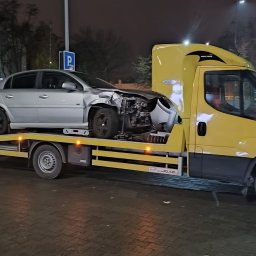 POMOC DROGOWA ŁUKASZ PRZYBORSKI - Żółta laweta z uszkodzonym srebrnym autem osobowym na platformie, nocne ujęcie na parkingu, widoczny znak parkingu.