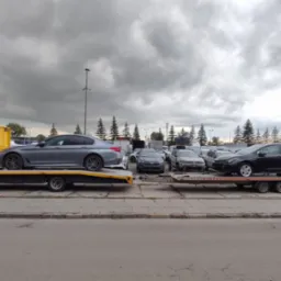 Laweta z szarym BMW na żółtej platformie, obok druga laweta z czarnym autem. W tle zaparkowane samochody i pochmurne niebo.