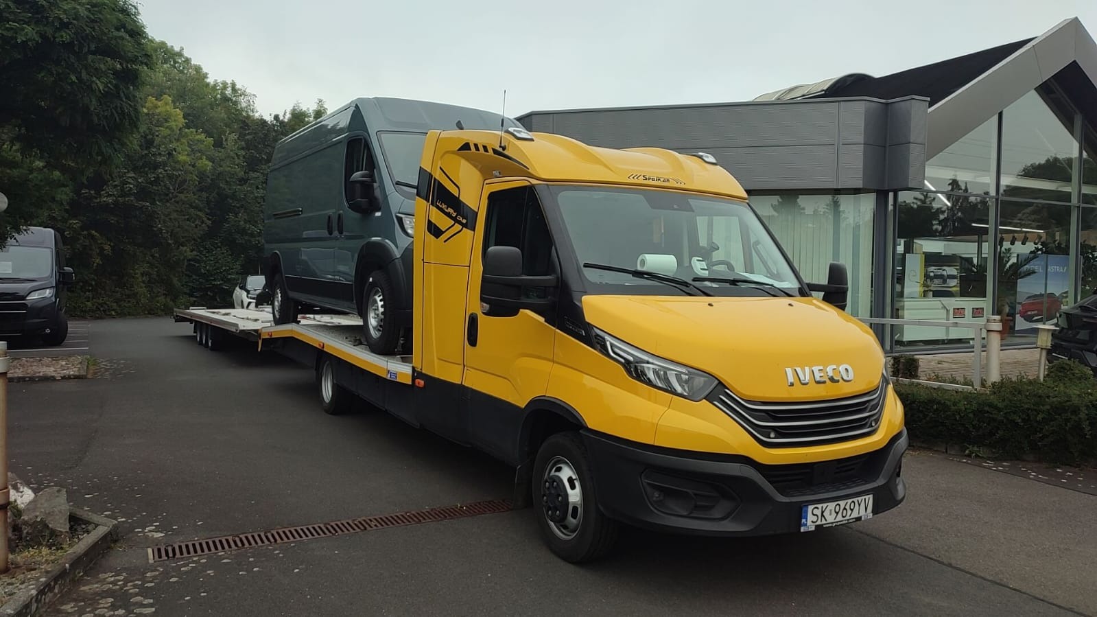Żółta laweta Iveco z szarym vanem na platformie, zaparkowana w Idzbark. Widoczny budynek z dużymi oknami w tle. Tablica rejestracyjna SK-969YV.