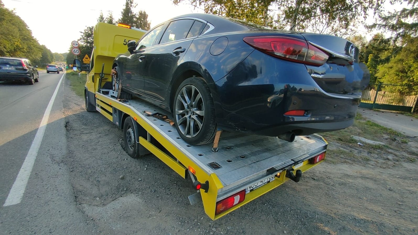 Żółta laweta z załadowanym, ciemnym samochodem osobowym marki Mazda na poboczu drogi, w tle zieleń drzew i inne pojazdy.