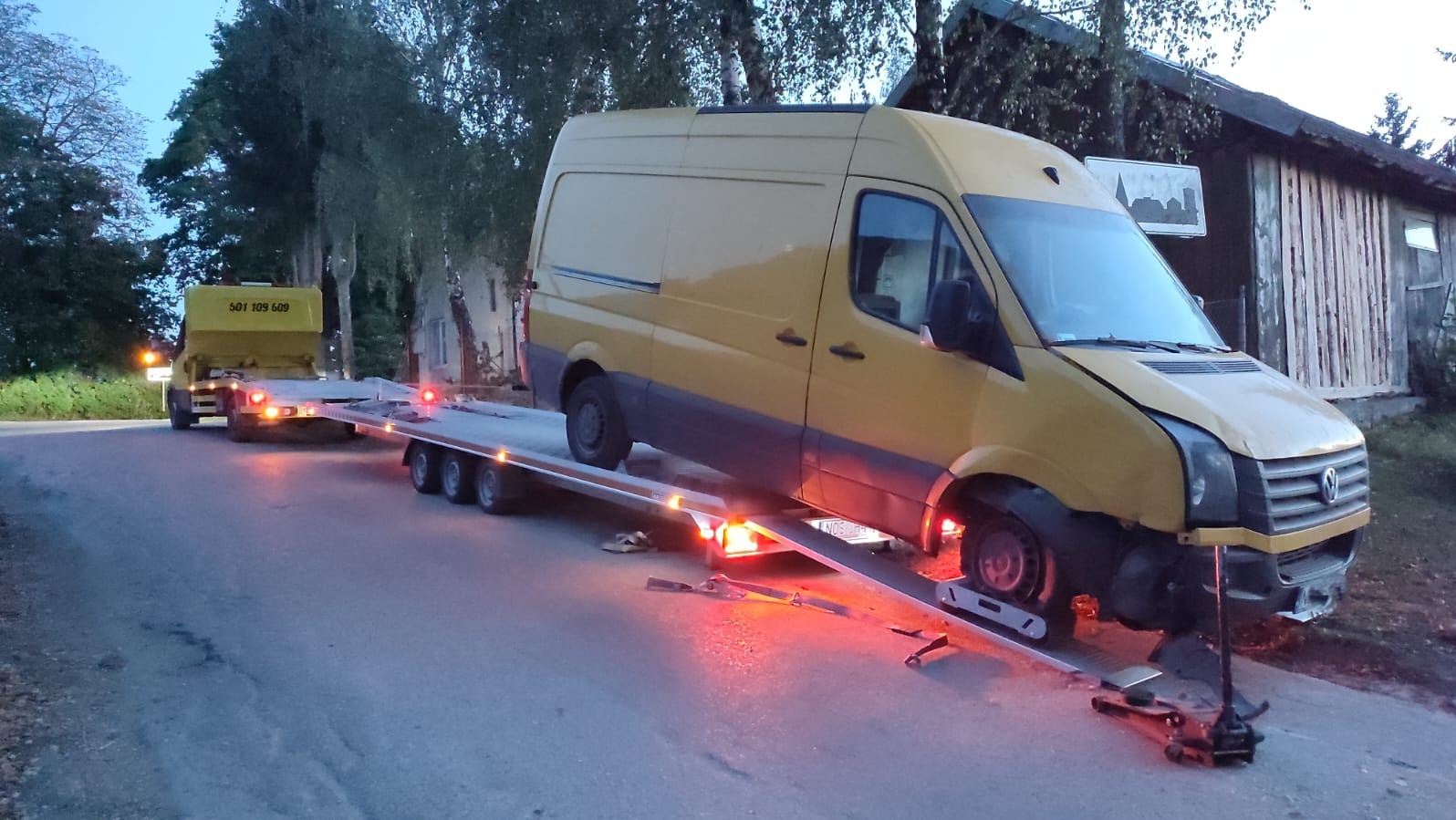 Żółty uszkodzony bus na lawecie z włączonymi światłami awaryjnymi, zaparkowany na poboczu drogi w pobliżu zalesionego terenu i drewnianego budynku.