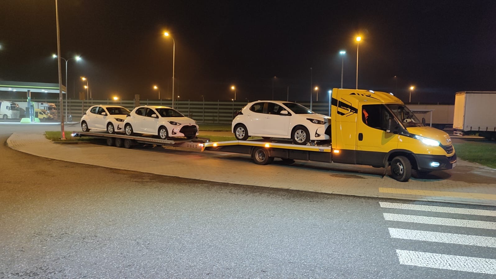 Żółta laweta z trzema białymi samochodami Toyota Yaris na przyczepie, nocą na parkingu stacji benzynowej, widok z boku.