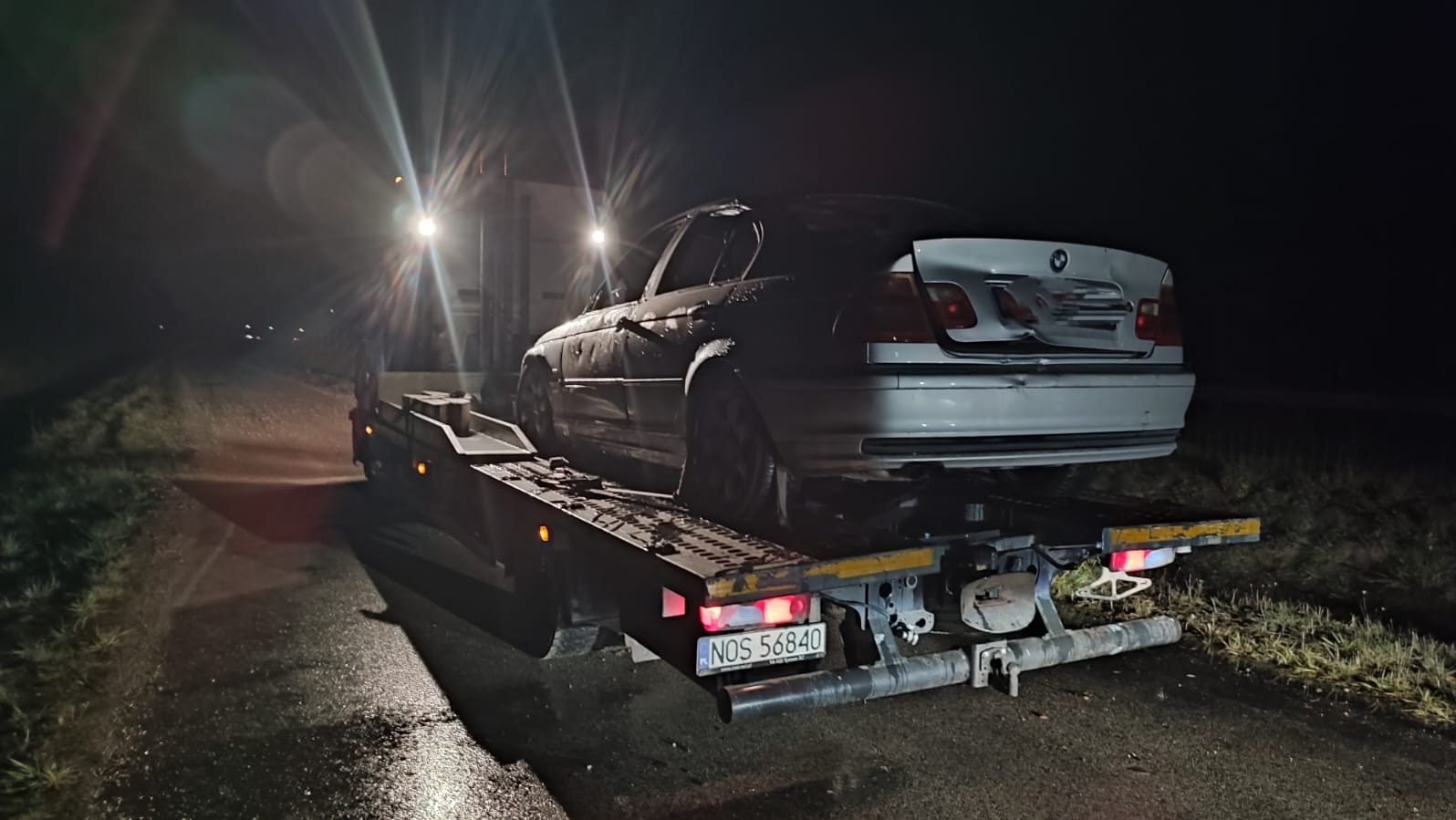 Uszkodzony, srebrny samochód BMW na platformie lawety w nocy. Widoczne uszkodzenia tylnej części pojazdu i oświetlenie lawety. Tablica rejestracyjna NOS 56840.