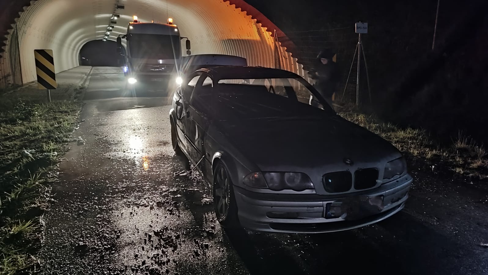Uszkodzone auto BMW z otwartą maską, holowane przez lawetę w tunelu. Mokra nawierzchnia, nocne oświetlenie, widoczny znak drogowy.