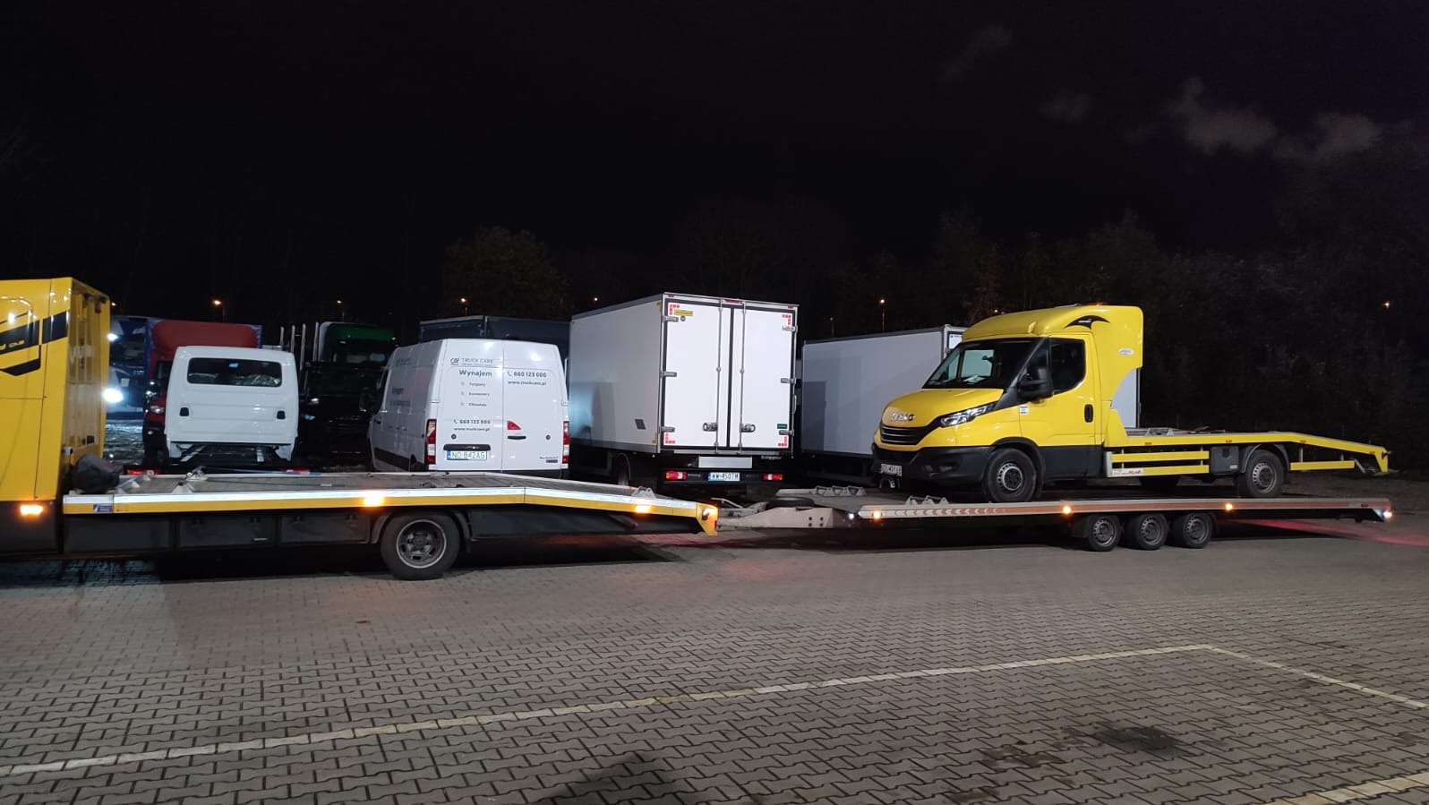 Żółta laweta Iveco z platformą załadowaną białym furgonem na parkingu nocą. W tle inne pojazdy dostawcze i ciężarowe.