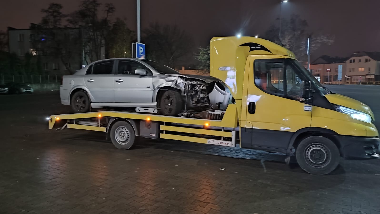 Żółta laweta z uszkodzonym srebrnym autem osobowym na platformie, nocne ujęcie na parkingu, widoczny znak parkingu.