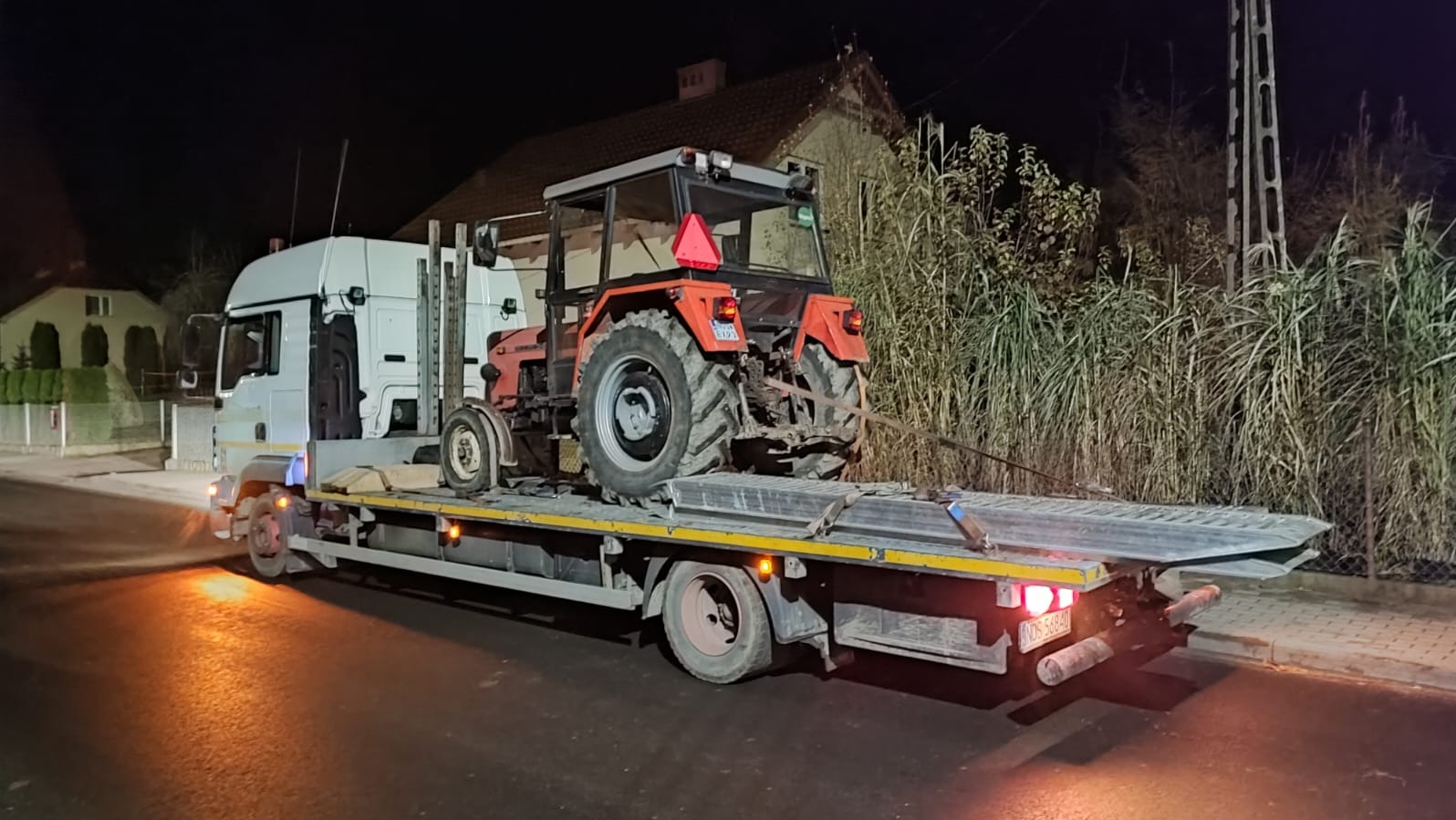 Laweta przewozi czerwony traktor rolniczy po zmroku. Widoczny fragment budynku w tle. Tylne światła lawety oświetlają jezdnię.