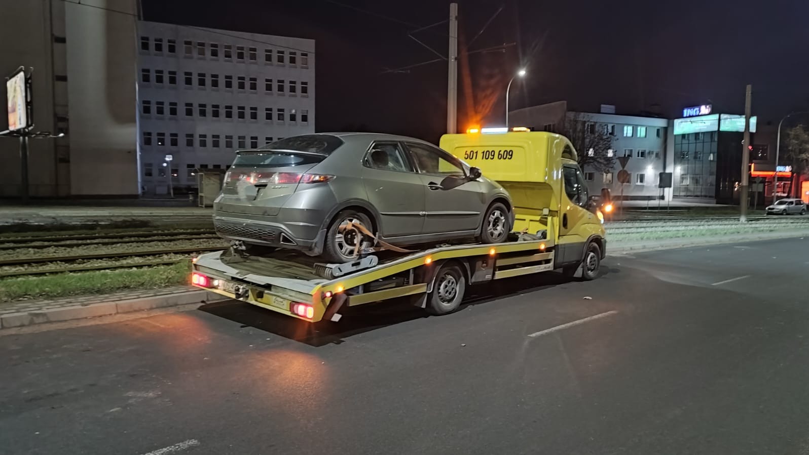 Szara Honda Civic na żółtej lawecie, zabezpieczona pasami transportowymi, na tle miejskiej scenerii nocą z budynkami i torami tramwajowymi.
