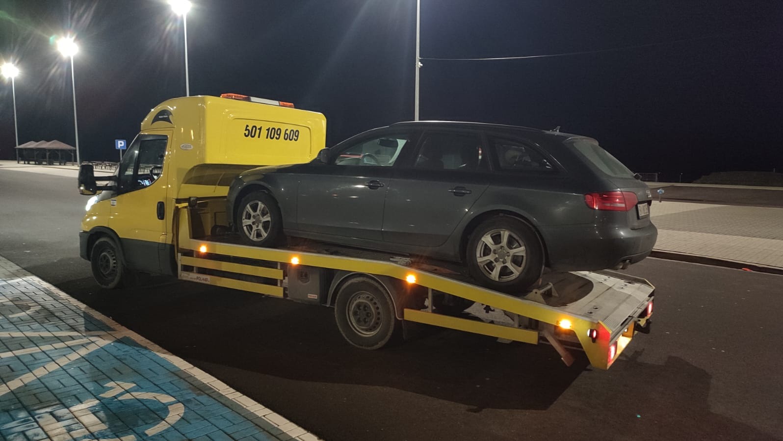 Żółta laweta z szarym kombi Audi na platformie, nocne oświetlenie uliczne, widoczny numer telefonu na kabinie lawety.