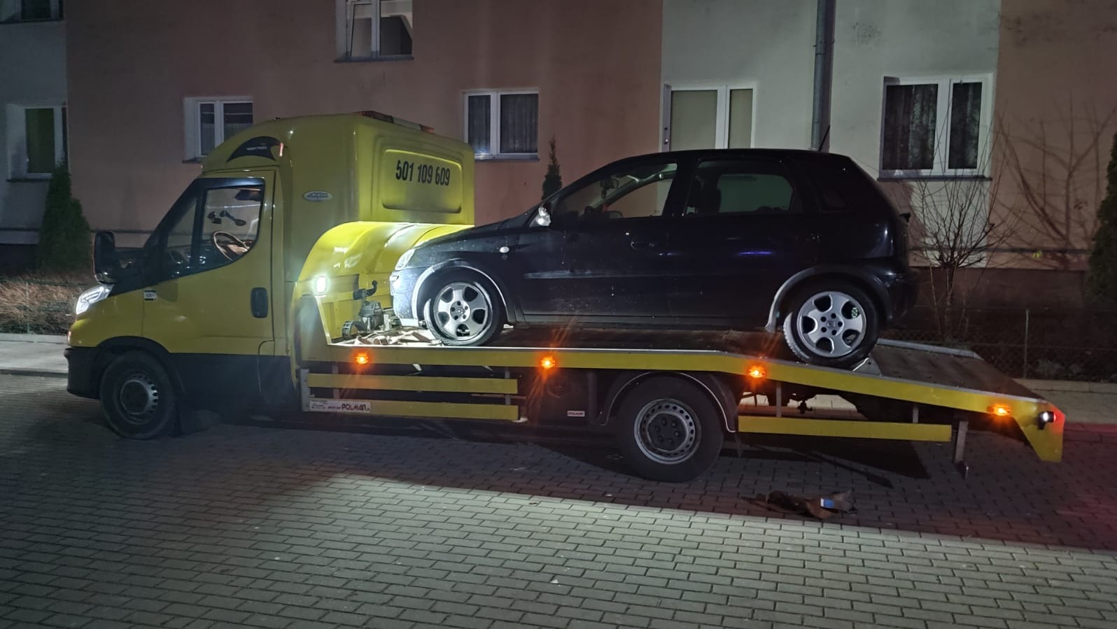 Żółta laweta z czarnym samochodem osobowym na platformie, zaparkowana na tle budynku mieszkalnego w nocnej scenerii. Widoczny numer telefonu na kabinie lawety.
