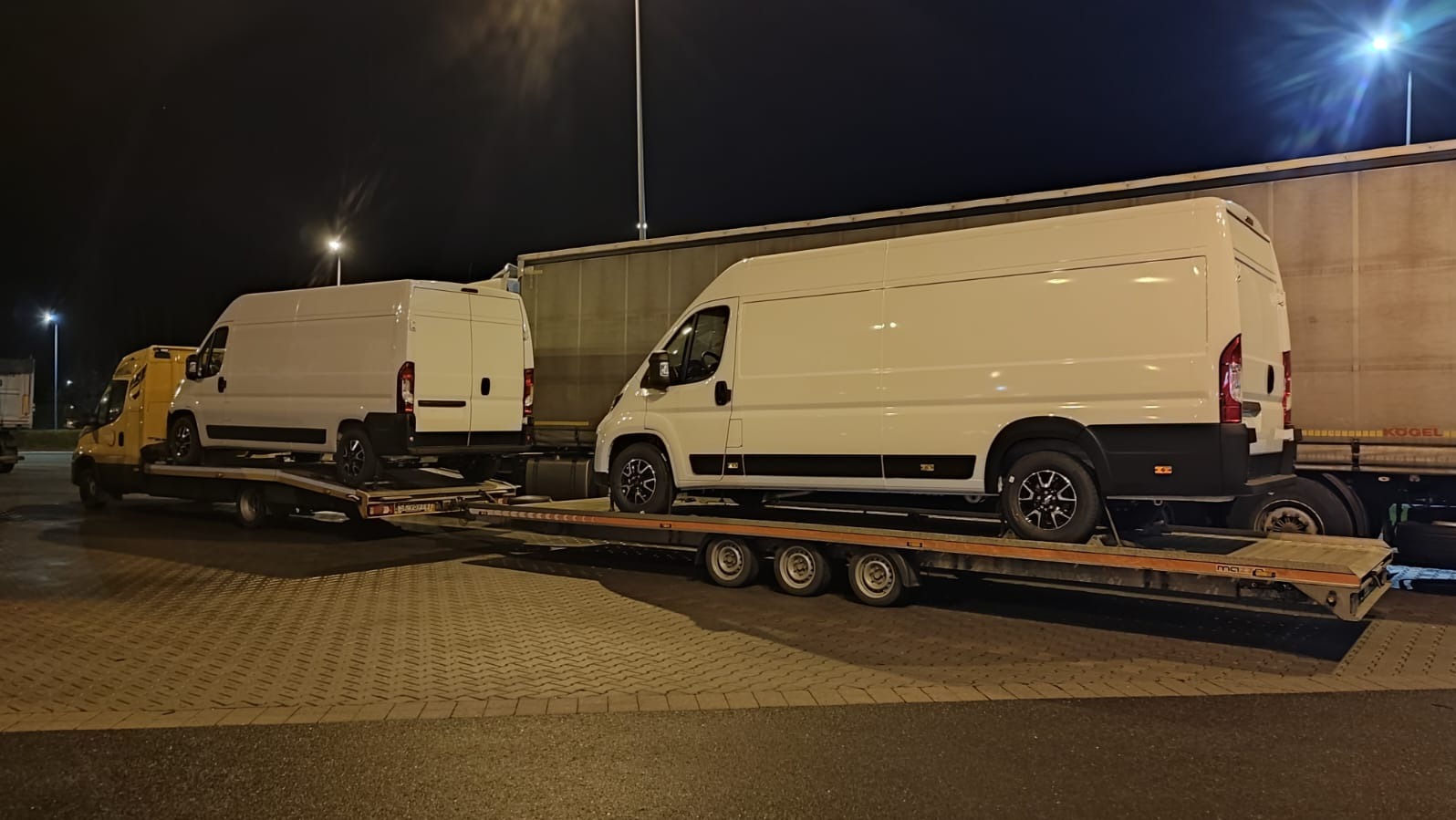Dwa białe busy na niskopodwoziowej lawecie, przewożone nocą. Z przodu żółty holownik, w tle naczepa ciężarówki z logo Kogel. Widoczny bruk i słupy oświetleniowe.