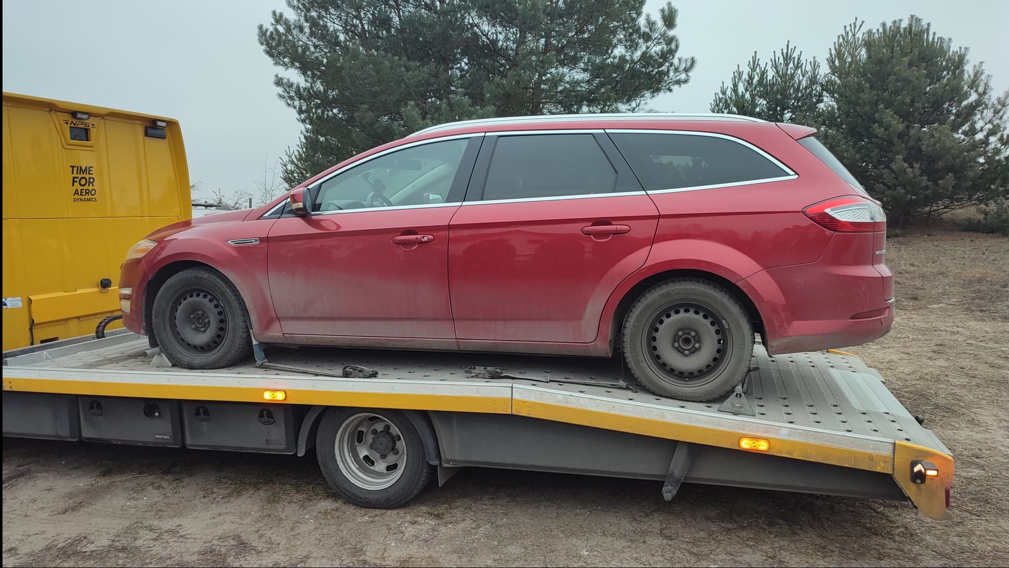 Czerwony Ford Mondeo kombi przewożony na żółtej lawecie w pochmurny dzień. Widoczne drzewa w tle. Auto lekko zabrudzone.
