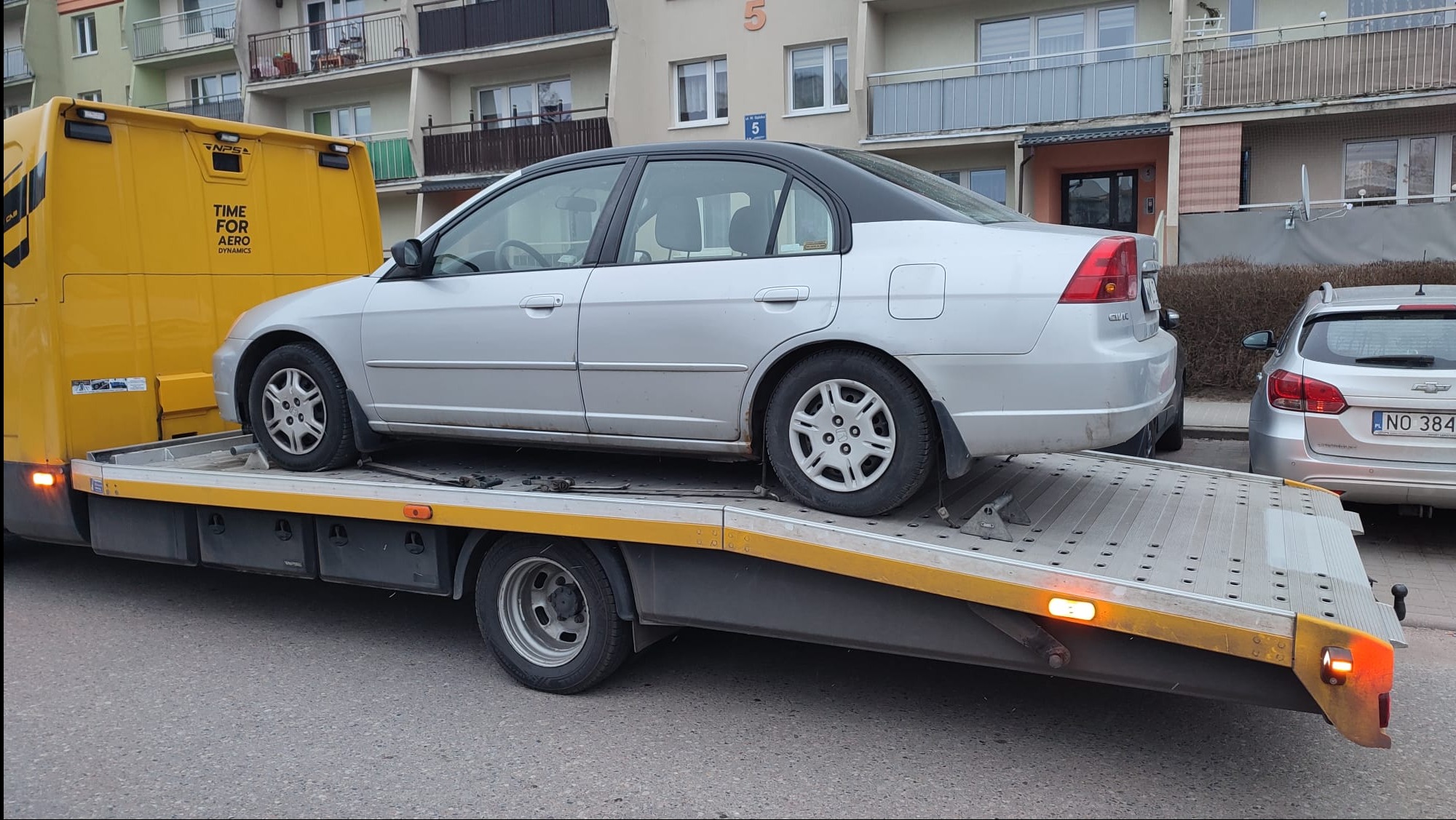 Srebrny samochód osobowy na żółtej lawecie, zaparkowanej na tle bloku mieszkalnego. Widoczny fragment innego auta z polską tablicą rejestracyjną.