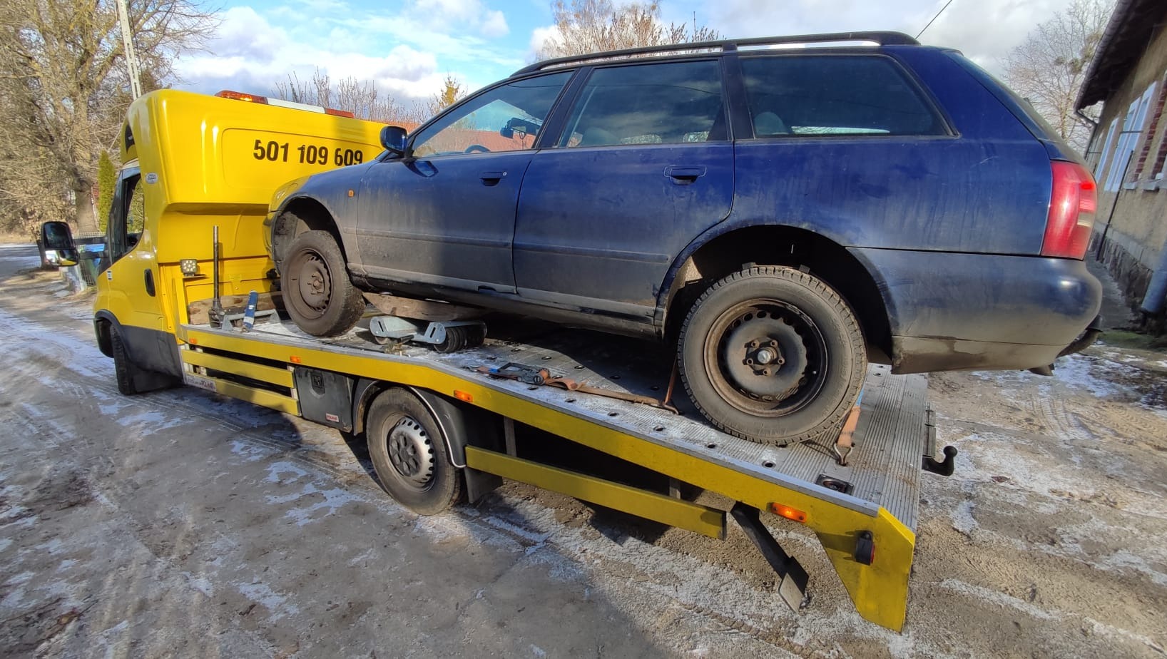 Niebieskie kombi na żółtej lawecie z numerem 501 109 609, na tle drzew i budynku. Laweta na drodze, częściowo pokrytej śniegiem. Słoneczny dzień.