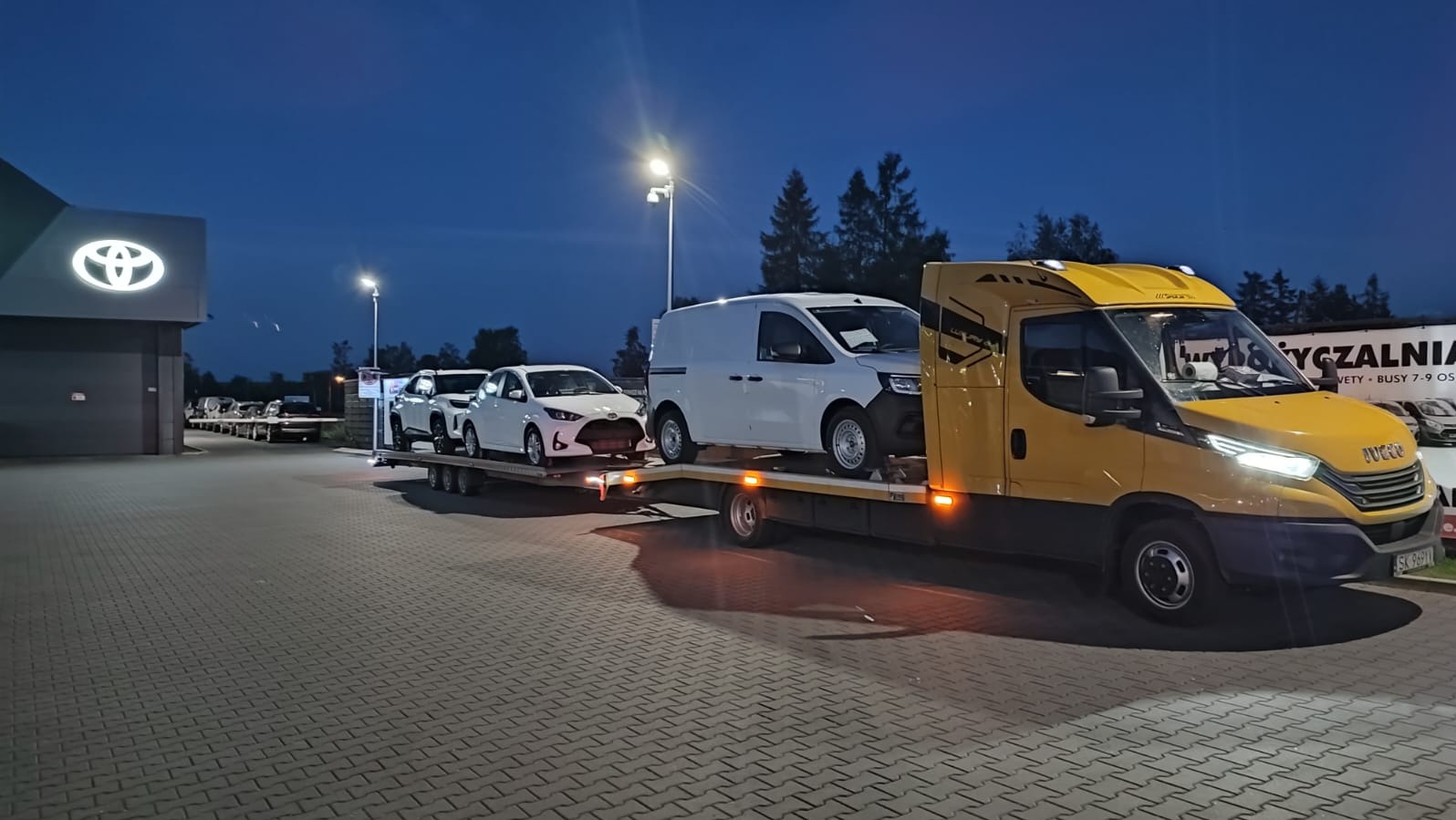 Żółta laweta Iveco z trzema białymi pojazdami Toyota na platformie, zaparkowana przed salonem samochodowym Toyota o zmierzchu.