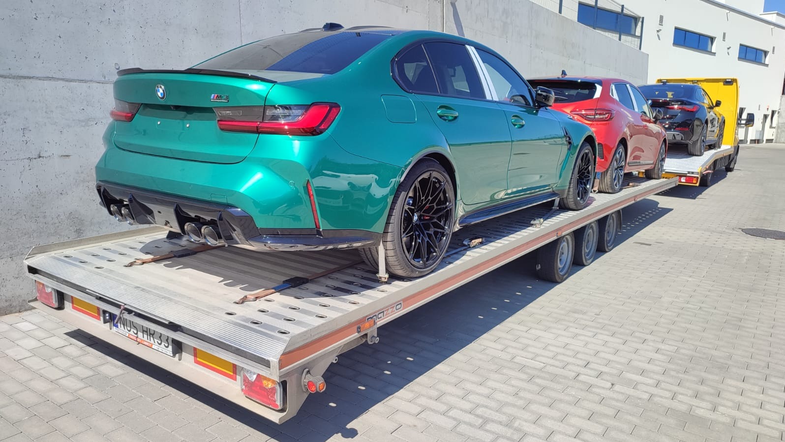 Laweta z trzema samochodami: zielonym BMW M3, czerwonym SUV-em i czarnym BMW. Widok z tyłu, na jasnym brukowanym placu.