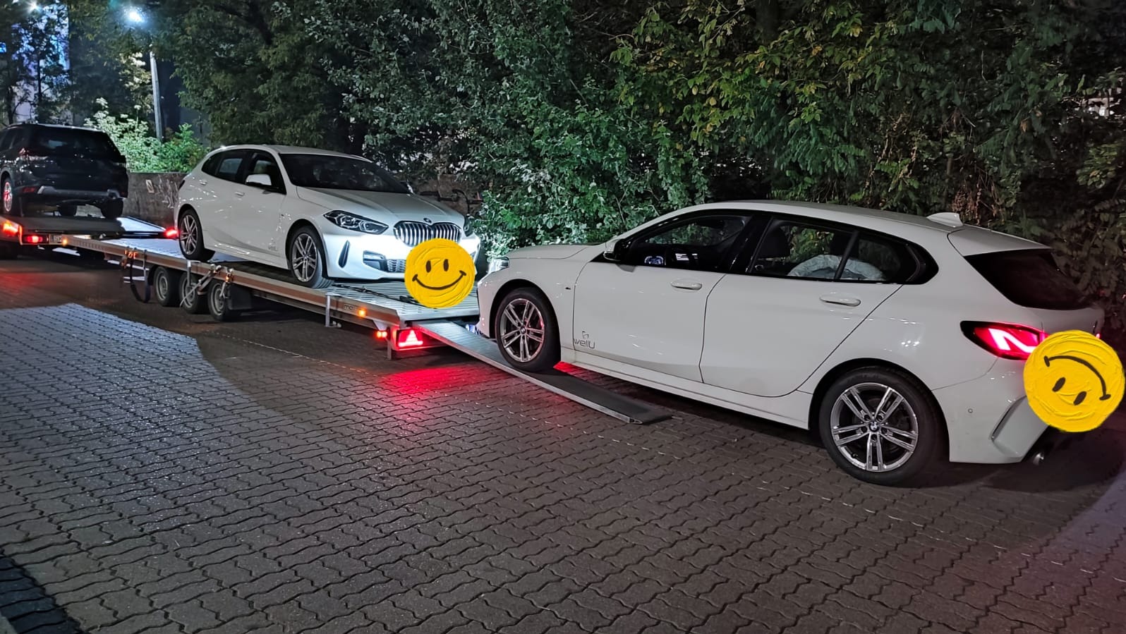 Laweta z dwoma białymi BMW na chodniku, jeden wjeżdża na platformę. Widoczne światła stopu i otoczenie z zielenią. Nocne ujęcie.