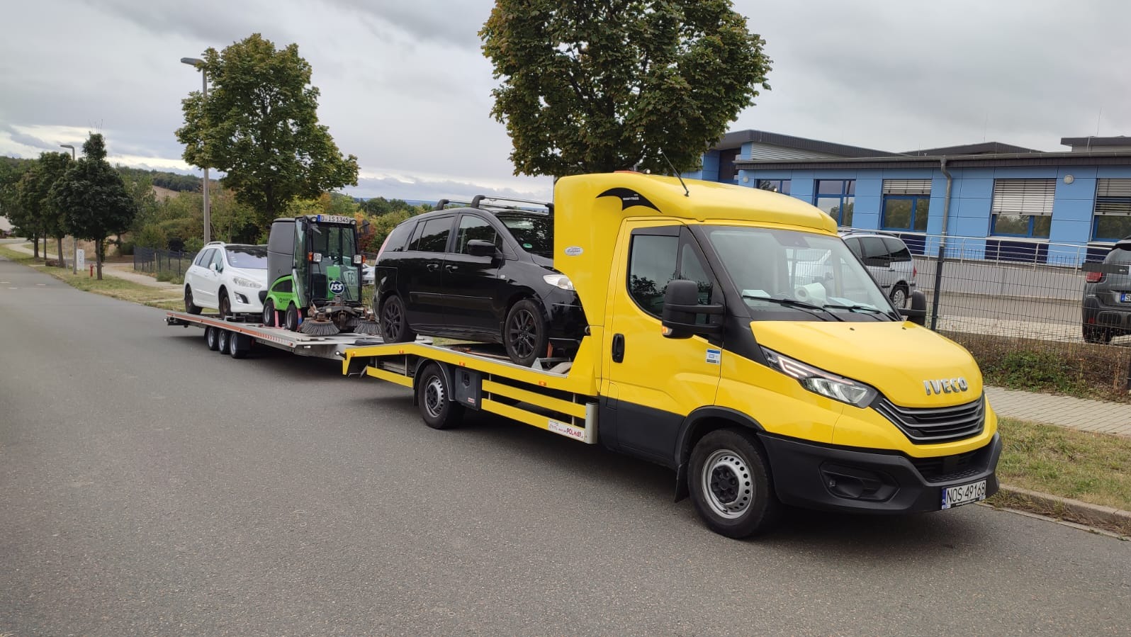 Żółta laweta Iveco z czarnym samochodem osobowym i zieloną maszyną czyszczącą na platformie, na tle szarej drogi i budynków.