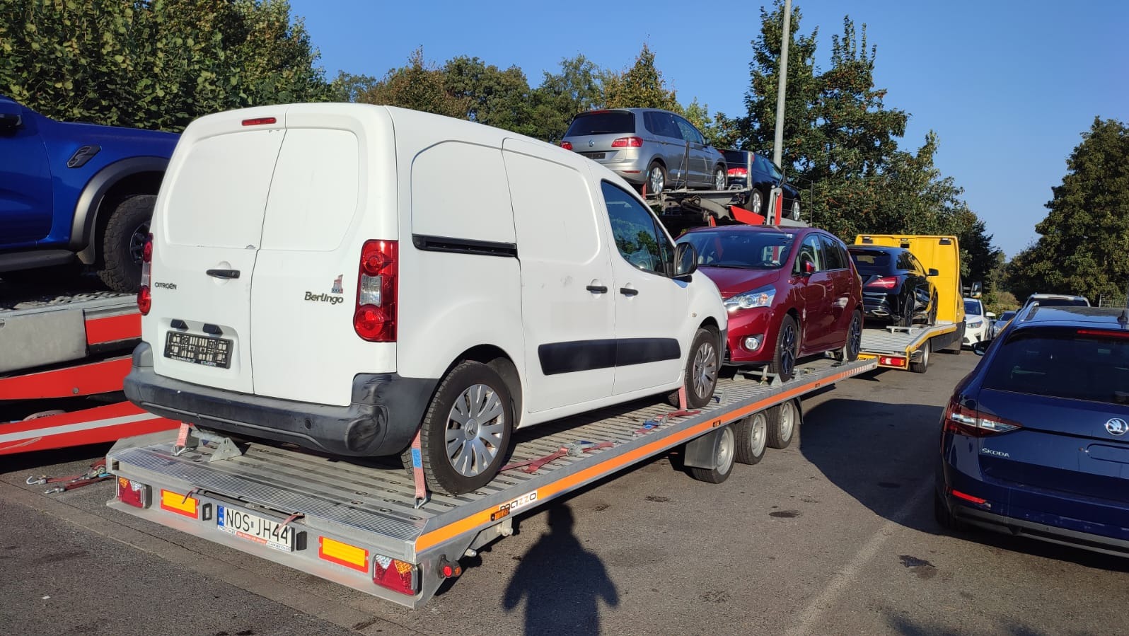 Laweta z różnymi autami: biały Citroen Berlingo, czerwony sedan, czarny Mercedes. Rejestracja lawety: NOS JH44. Widok z boku na załadowaną lawetę.