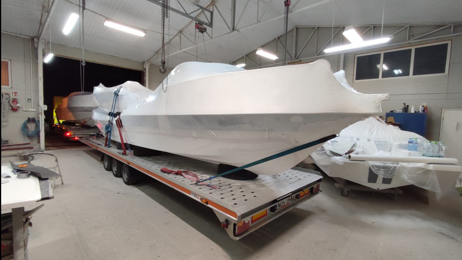 Łódź motorowa owinięta folią, zabezpieczona pasami, na niskopodwoziowej lawecie w hali. W tle widoczna druga łódź i fragment ciężarówki.