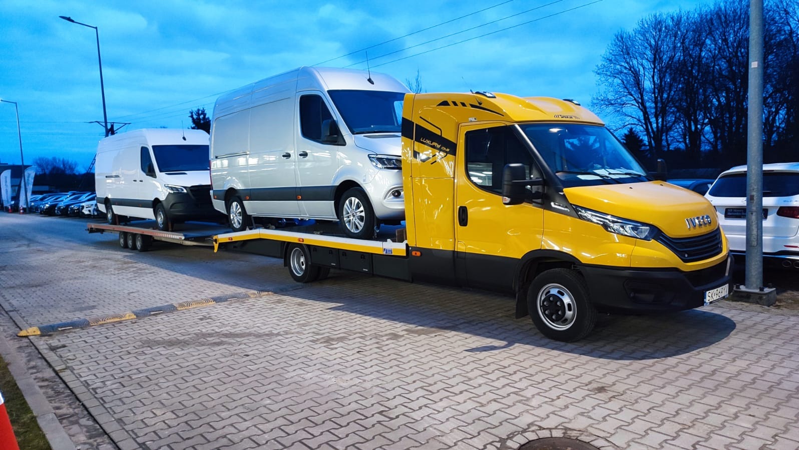 Żółta laweta Iveco przewozi dwa białe busy Mercedes Sprinter. Ujęcie z boku, widoczne detale pojazdów i otoczenie parkingu. Tablica rejestracyjna lawety widoczna.