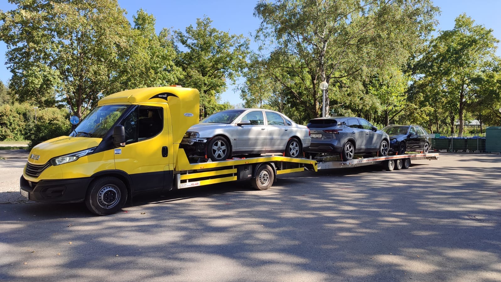 Żółta laweta z trzema samochodami osobowymi na platformie, zaparkowana na parkingu w słoneczny dzień. Widoczne drzewa w tle.