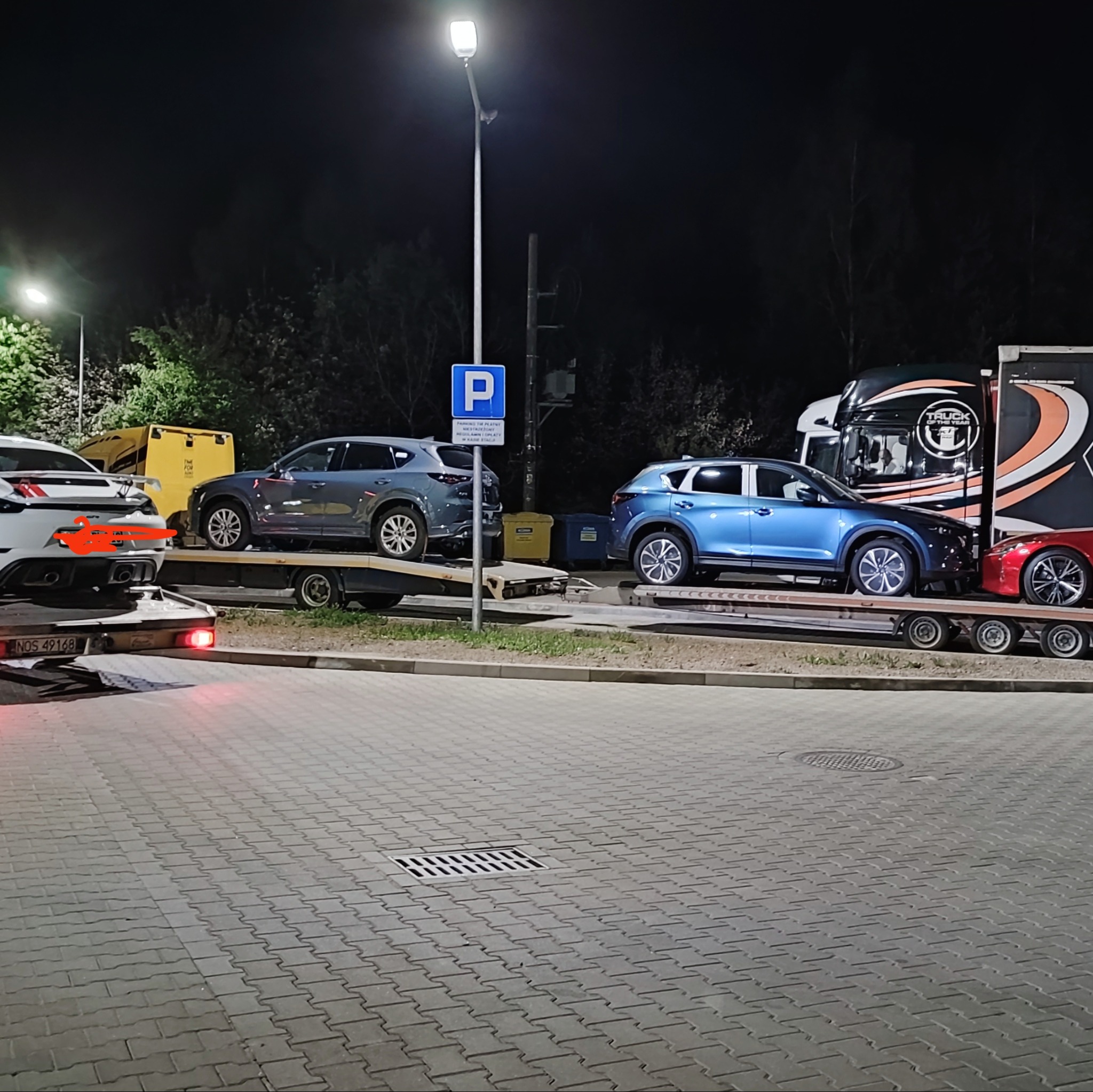 Nocny transport czterech samochodów osobowych na lawecie, oświetlonej światłem ulicznej latarni. Widoczny znak parkingu.