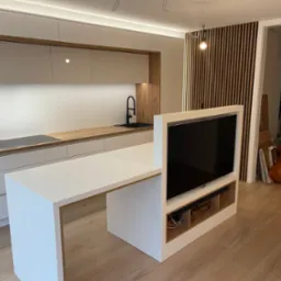 Nowoczesna kuchnia z wyspą, drewnianymi akcentami i telewizorem. Białe szafki, drewniany blat i panel ścienny. Minimalistyczny design i oświetlenie LED.