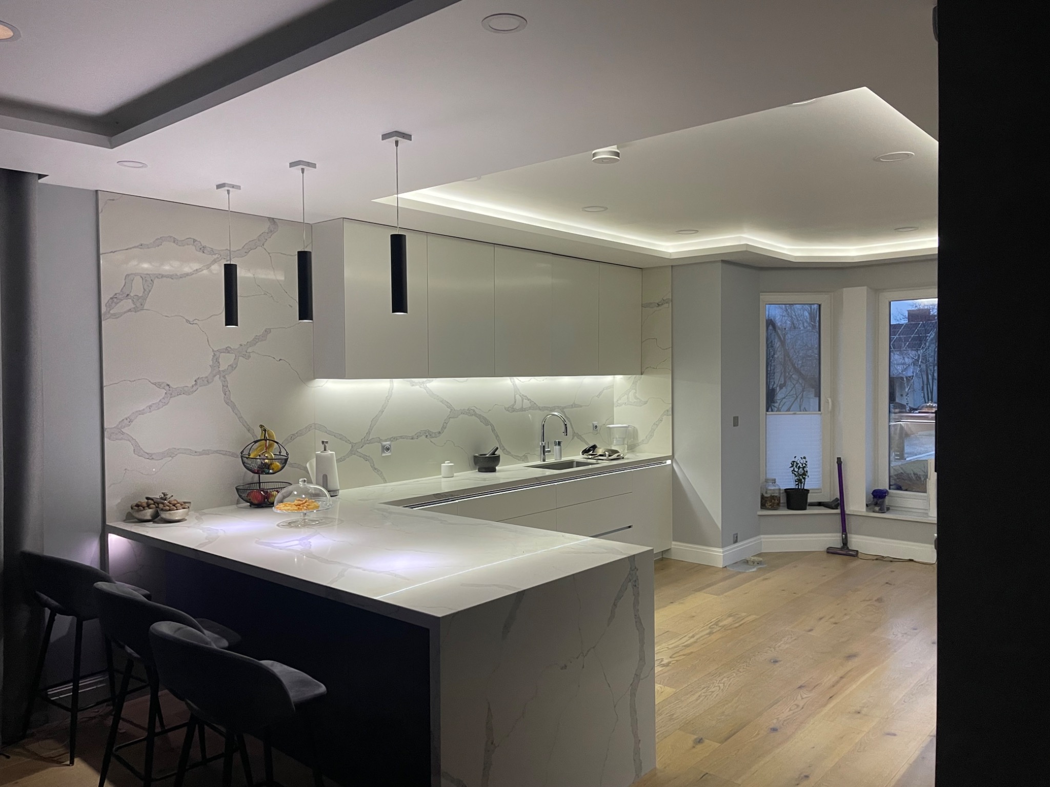 Nowoczesna kuchnia z wyspą i marmurowym blatem, oświetleniem LED pod szafkami i dekoracyjnymi lampami wiszącymi. Jasne wnętrze z drewnianą podłogą.