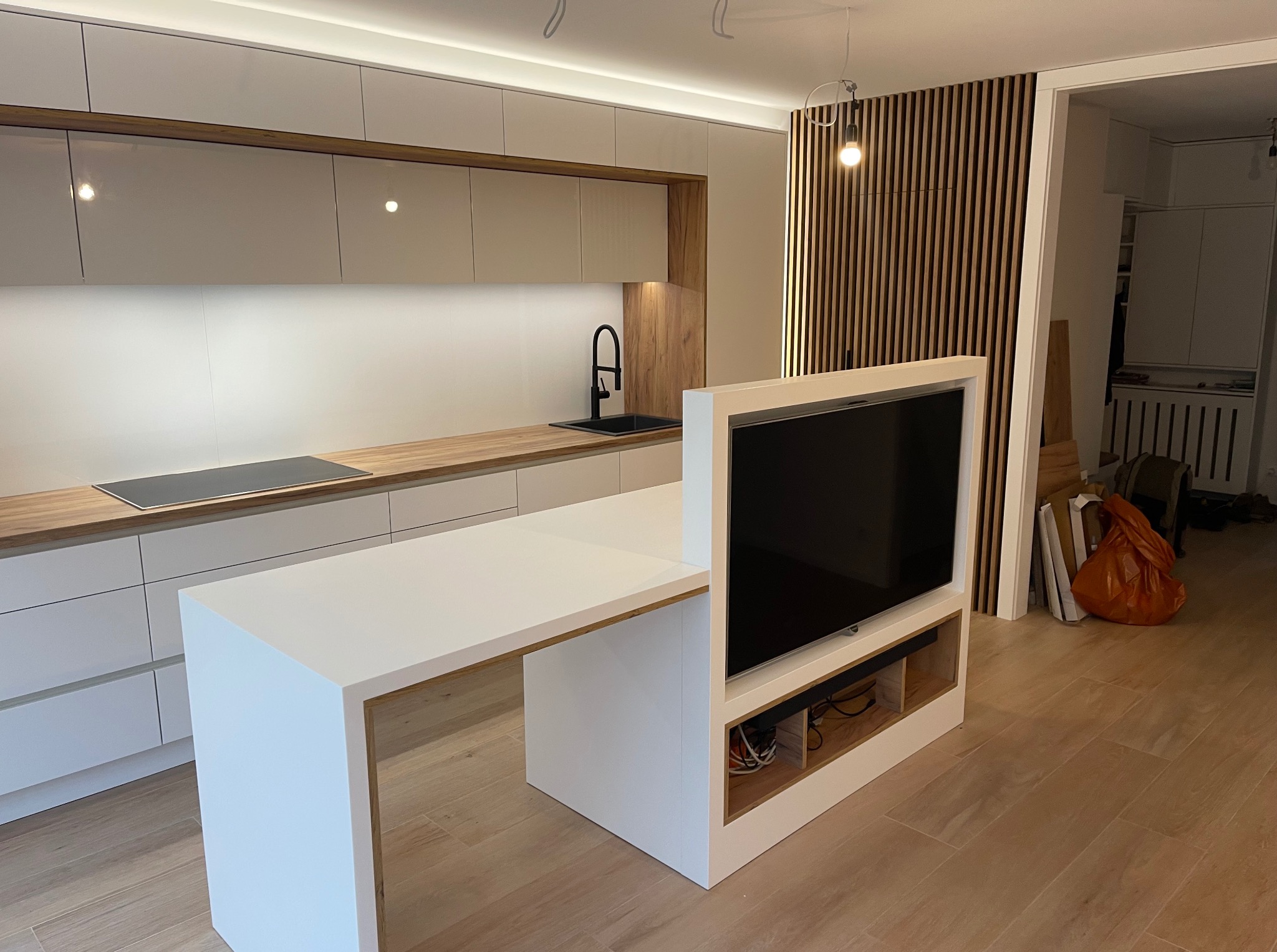 Nowoczesna kuchnia z wyspą, drewnianymi akcentami i telewizorem. Białe szafki, drewniany blat i panel ścienny. Minimalistyczny design i oświetlenie LED.