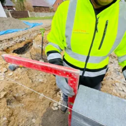 Pracownik w odblaskowej kurtce, z czerwoną poziomicą, układa dużą, szarą płytę. W tle wykop, ziemia, budynki. Widoczna łopata i biały sznurek wyznaczający poziom.