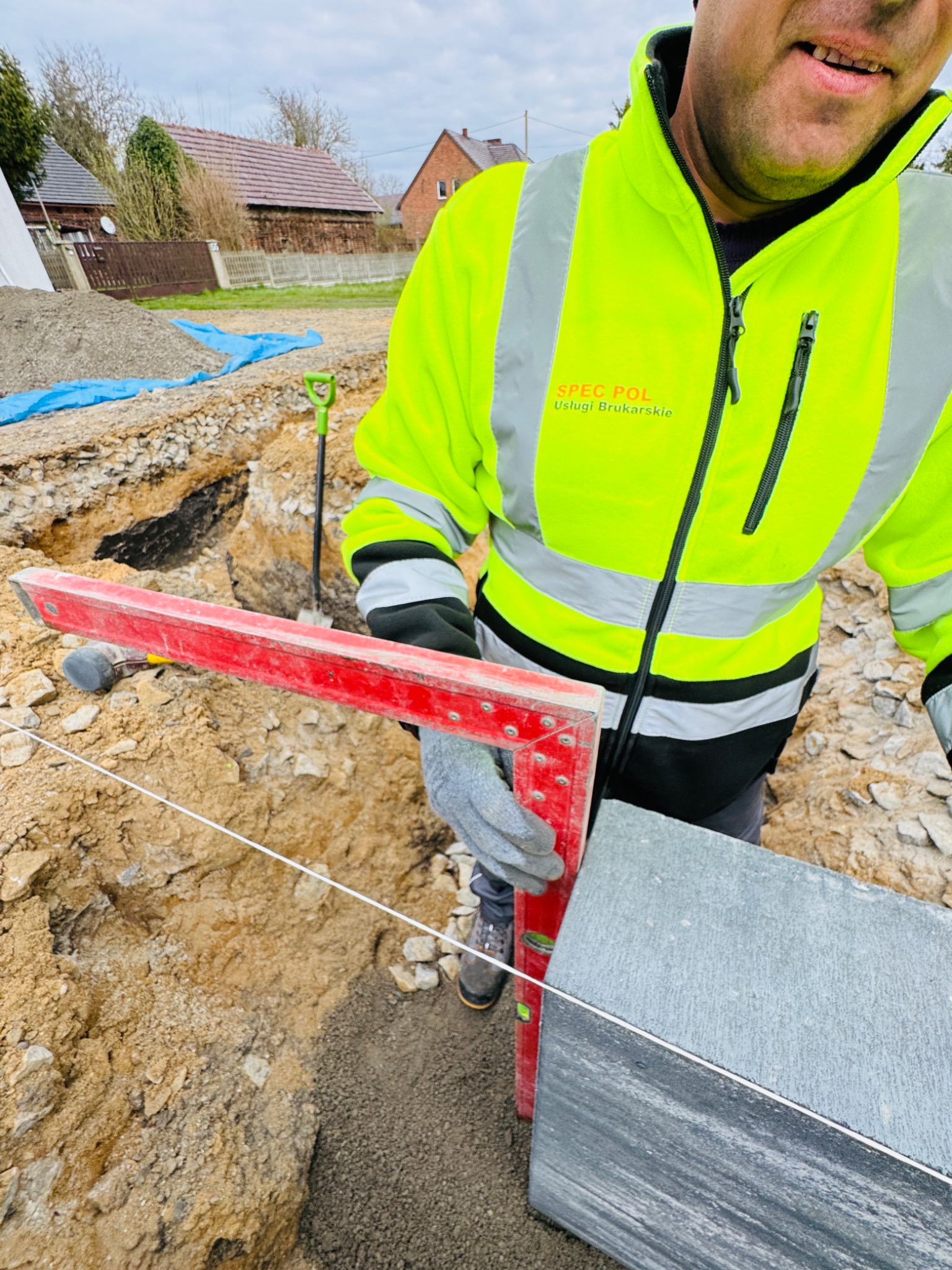 Pracownik w odblaskowej kurtce, z czerwoną poziomicą, układa dużą, szarą płytę. W tle wykop, ziemia, budynki. Widoczna łopata i biały sznurek wyznaczający poziom.