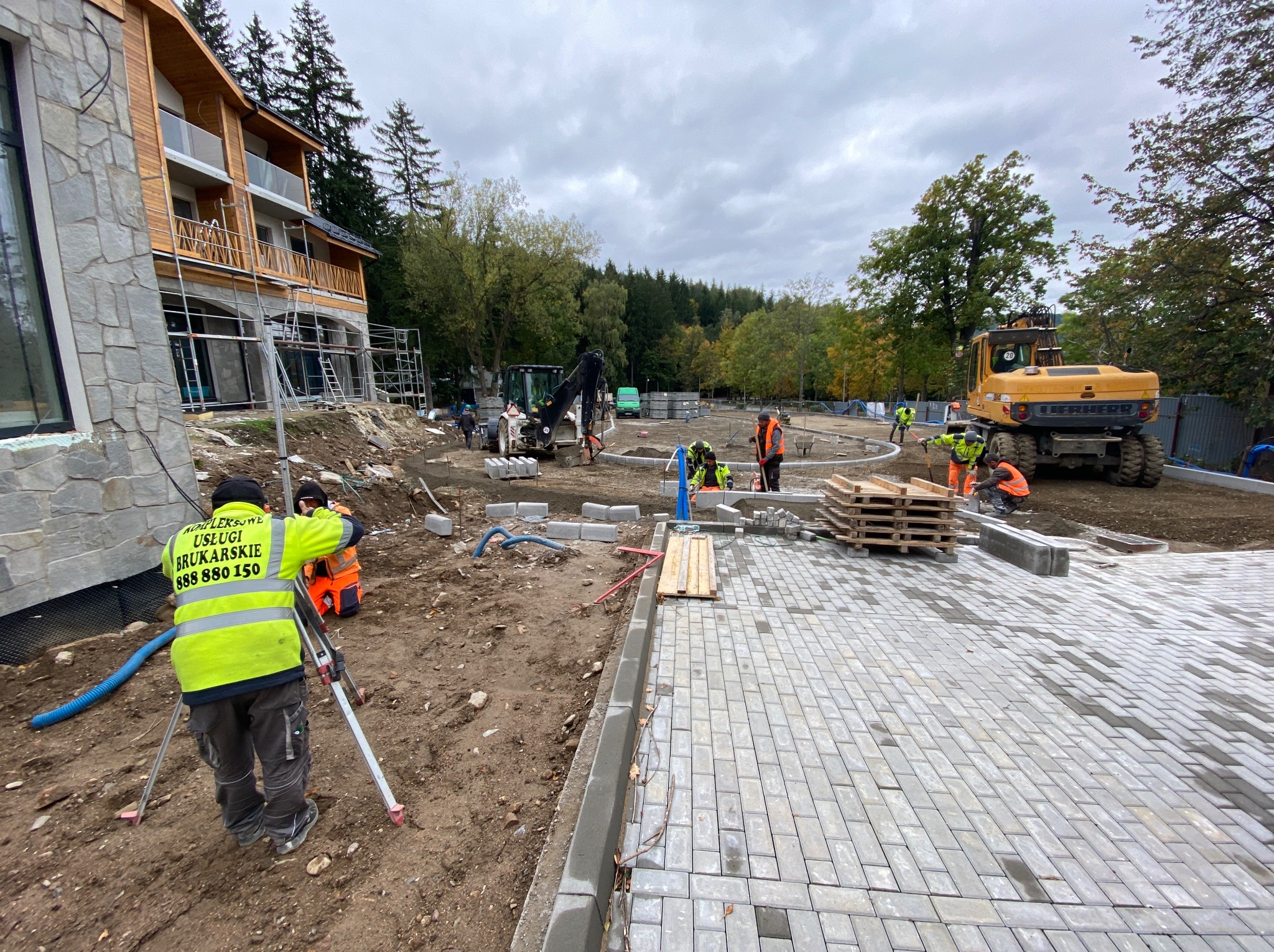 Ekipa brukarska w trakcie pracy: geodeta z niwelatorem, ułożona część kostki, palety z kostką, koparka na placu budowy. Widok na budynek w tle.