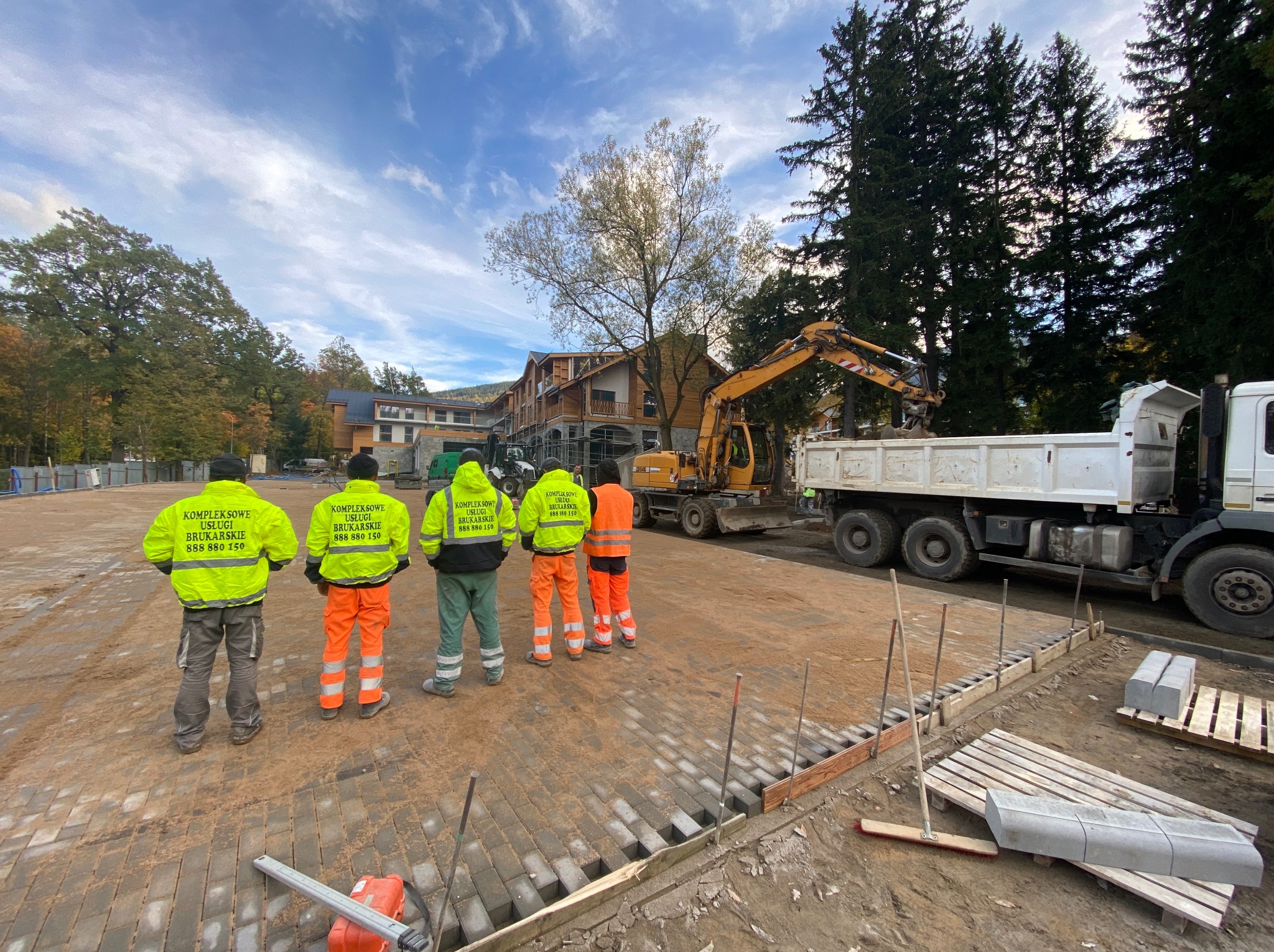 Ekipa brukarska w Opatowie na placu budowy, przygotowuje podłoże pod kostkę. W tle ciężarówka i koparka. Widoczne narzędzia i materiały budowlane.