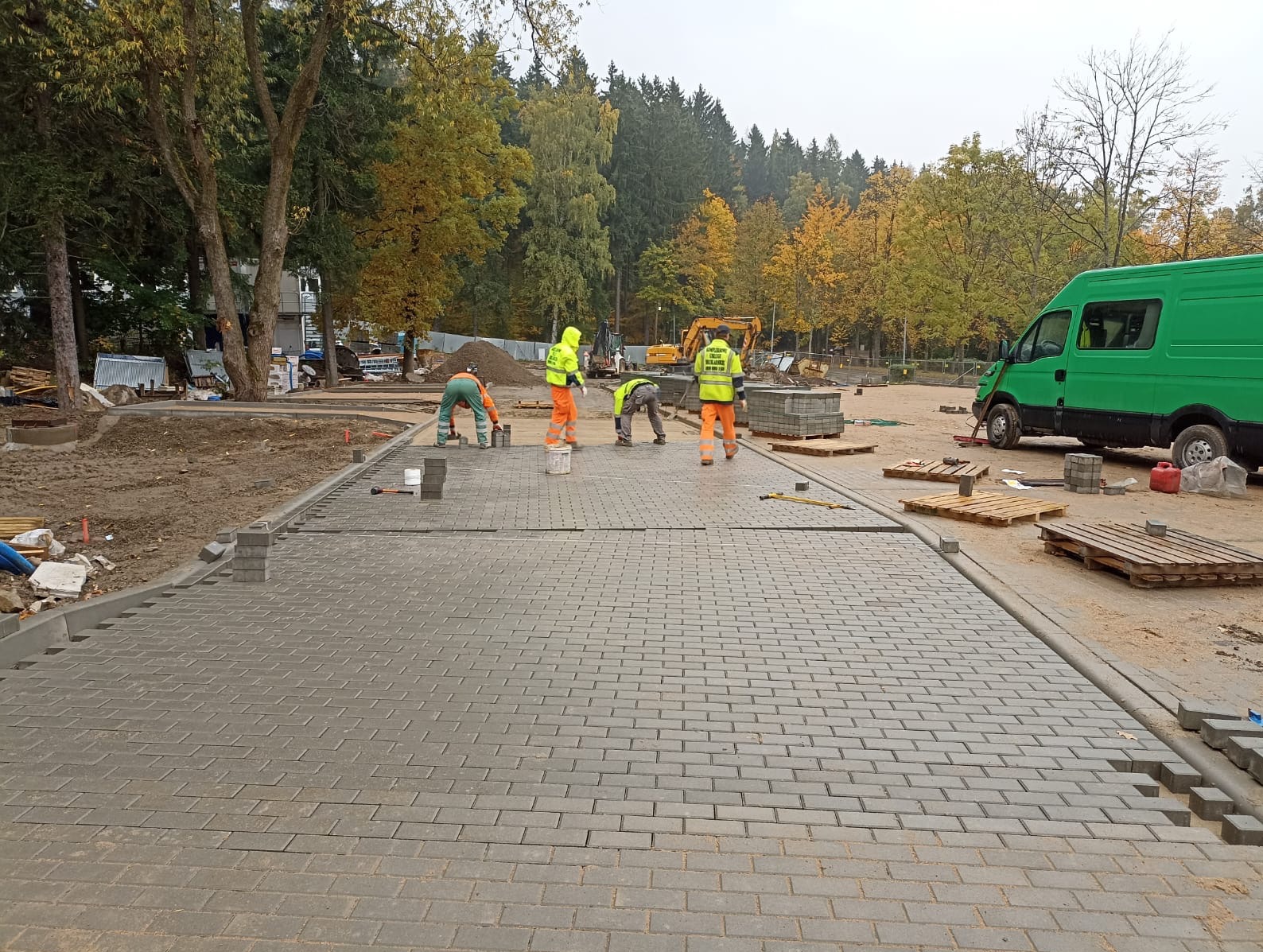Brygada w jaskrawych kamizelkach układa szarą kostkę brukową na placu budowy. W tle drzewa z jesiennymi liśćmi i zielony bus. Na placu budowy stoi koparka.