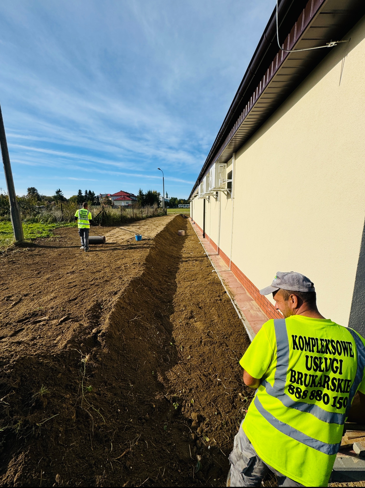 Przygotowanie terenu pod kostkę brukową: wykopany rów wzdłuż budynku, pracownicy w odblaskowych kamizelkach, wałek do ubijania ziemi w tle. Błękitne niebo.