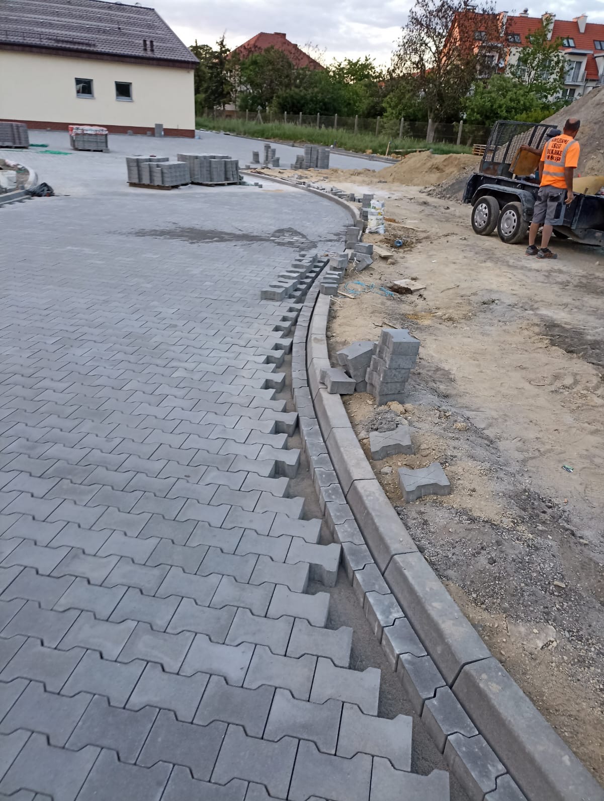 Etap układania szarej kostki brukowej typu I na posesji, widoczne obrzeża i przygotowane palety kostki. Pracownik w odblaskowej kamizelce przyczepia przyczepkę.