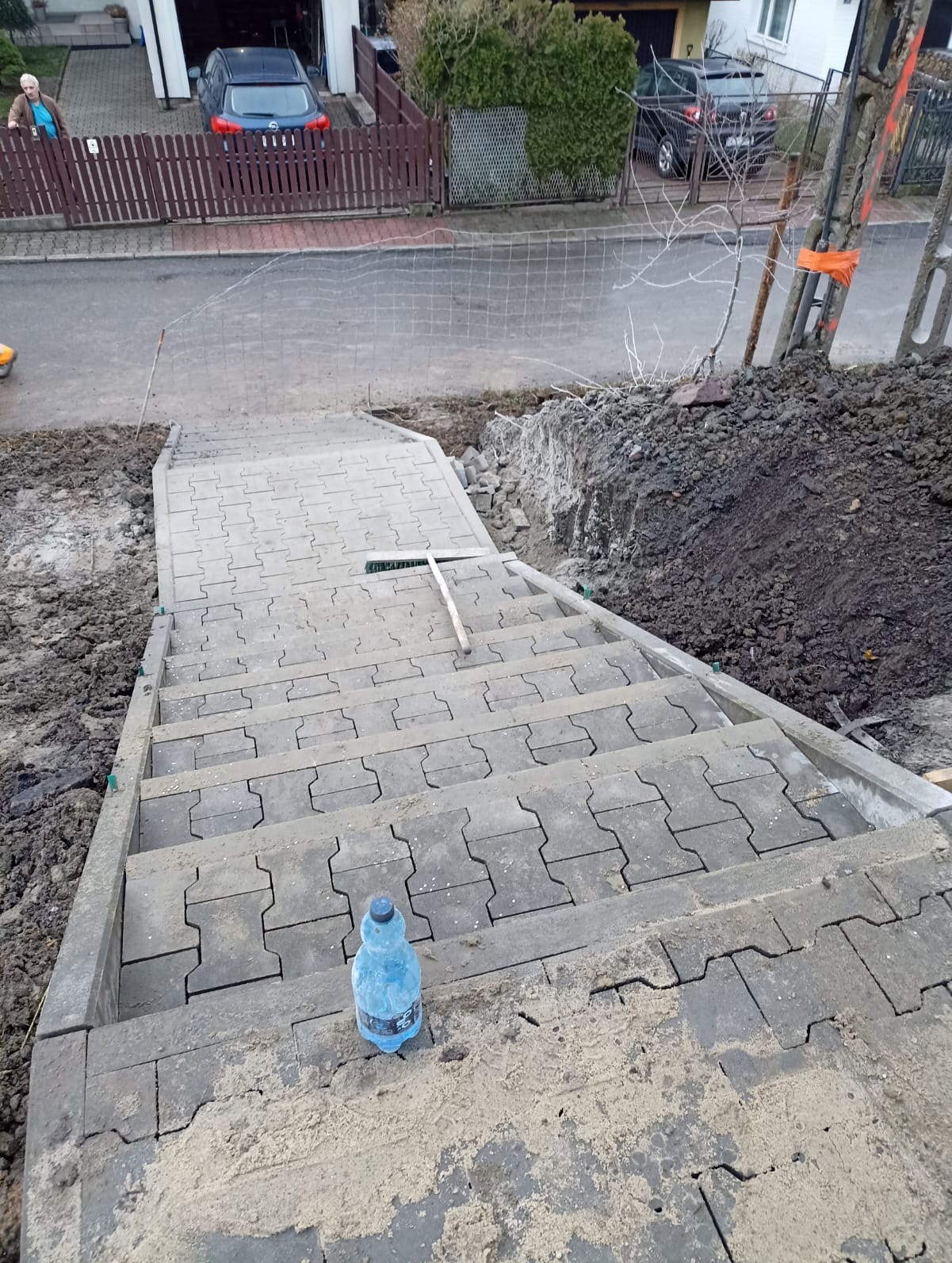 Schody z kostki brukowej w trakcie budowy, widok z góry, z butelką wody na dole i szczotką na jednym ze stopni. W tle ulica i domy.