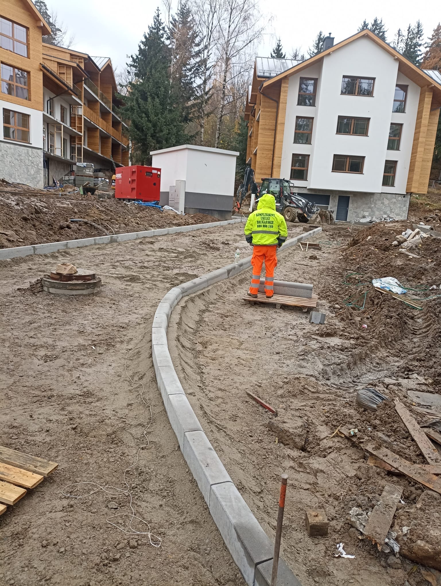 Pracownik w odblaskowym ubraniu układa obrzeża chodnika z szarej kostki betonowej na placu budowy. W tle budynki i sprzęt budowlany.