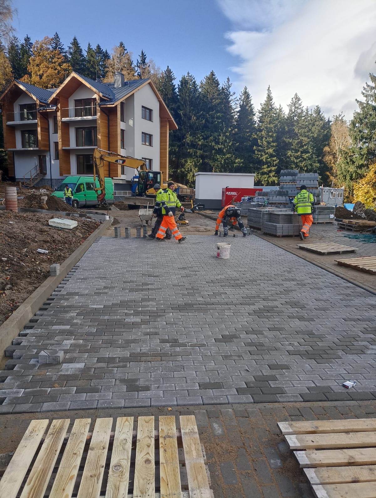 Ekipa w odblaskowych strojach układa szarą kostkę brukową na podjeździe. W tle budynek z balkonami i koparka CAT.