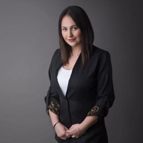 Portret kobiety w czarnej marynarce ze wzorem panterki na rękawach, na szarym tle. Uśmiechnięta, z ciemnymi włosami do ramion. Profesjonalny wygląd.