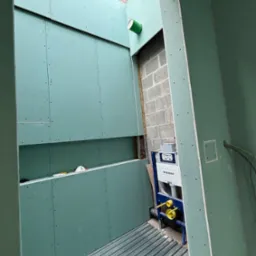 Wnętrze łazienki w trakcie montażu płyt gipsowo-kartonowych, widoczny stelaż podtynkowy Geberit i fragment instalacji. Ściany w kolorze zielonym, surowa cegła w tle.