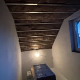 Skośny sufit poddasza z izolacją z wełny mineralnej i metalowym stelażem, widok z dołu, okno w tle. Część materiał&oacute;w budowlanych na dole kadru.