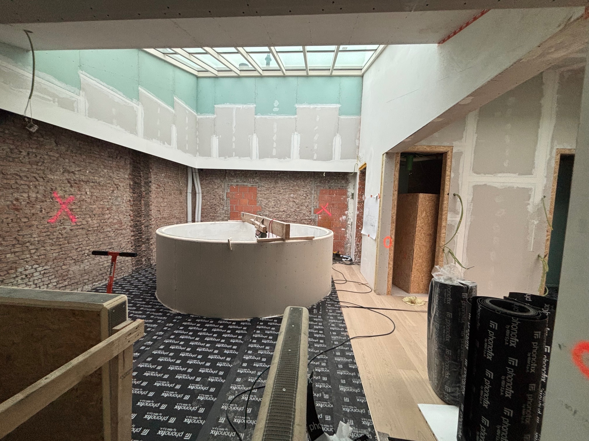 Wnętrze w trakcie prac: surowe ściany z cegły i płyty G-K, okrągła konstrukcja z płyt, materiały izolacyjne na podłodze, świetlik w suficie, widoczne instalacje.