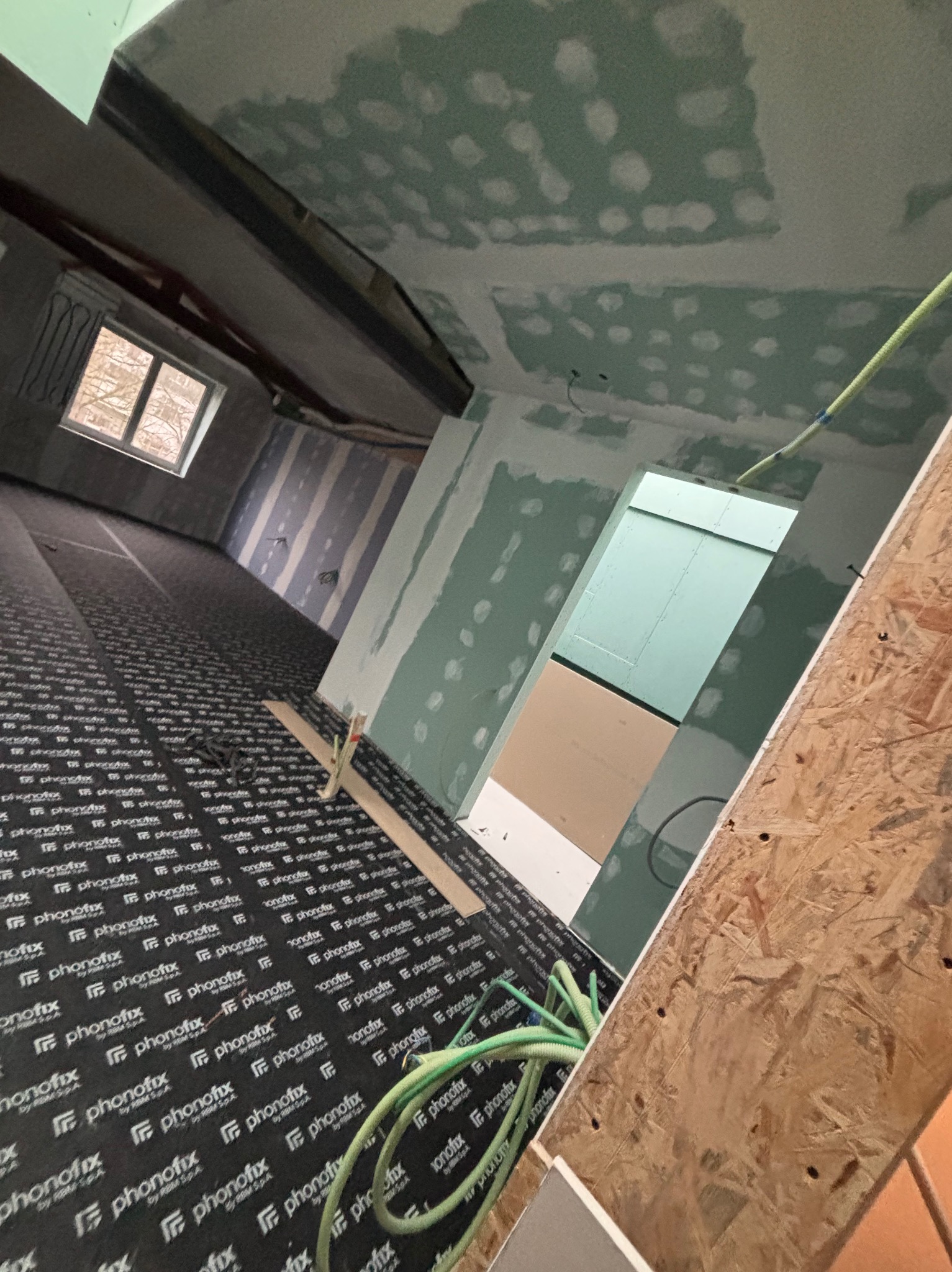 Wnętrze w trakcie prac wykończeniowych: położona izolacja podłogowa, ściany przygotowane pod szpachlowanie, sufit z widocznymi łączeniami płyt gipsowych.