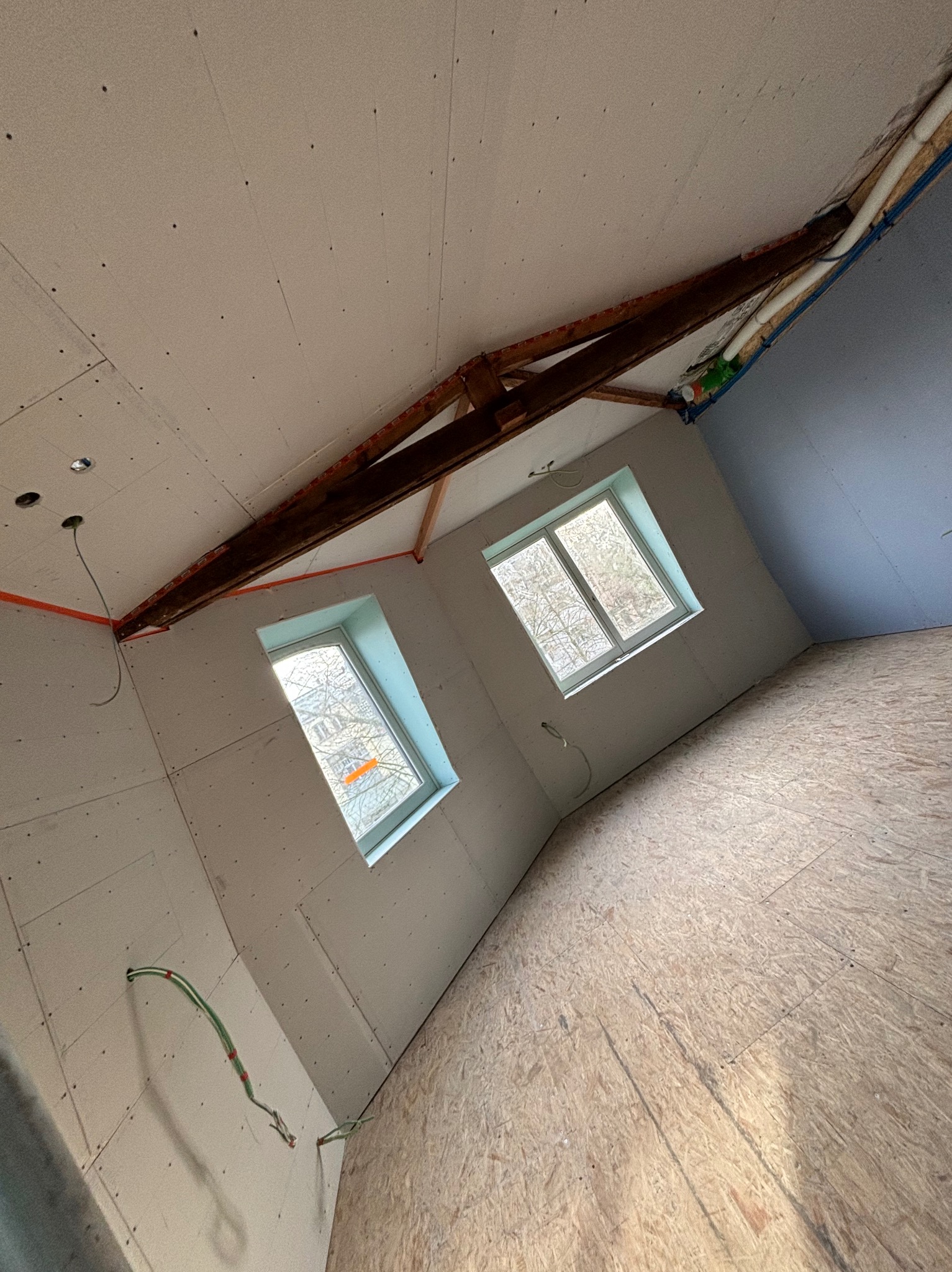 Poddasze w trakcie wykańczania: ściany i sufit pokryte płytami gipsowo-kartonowymi, widoczne okna dachowe i drewniana konstrukcja więźby dachowej. Częściowo położona podłoga z płyt OSB.