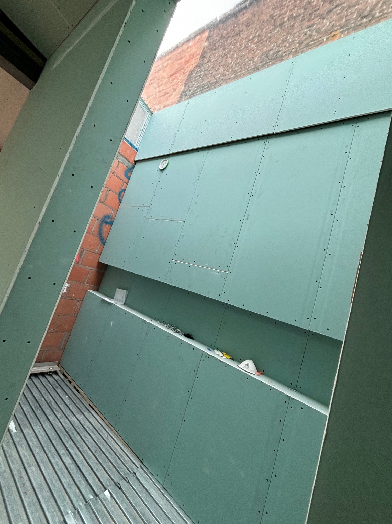 Zielone płyty gipsowo-kartonowe zamontowane na ścianie z cegły, widok z dołu. Widoczne ślady po wkrętach i niedokończone łączenia płyt.