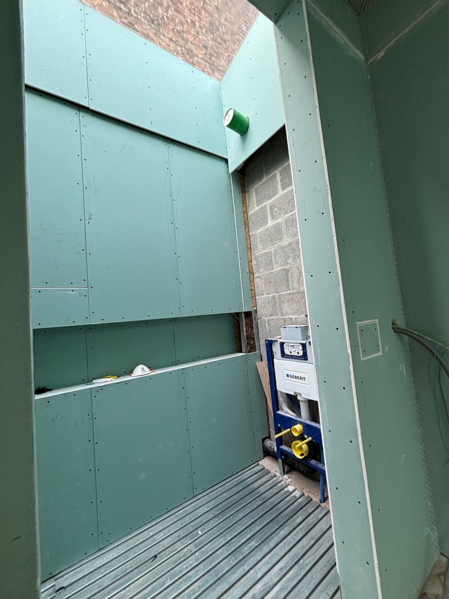 Wnętrze łazienki w trakcie montażu płyt gipsowo-kartonowych, widoczny stelaż podtynkowy Geberit i fragment instalacji. Ściany w kolorze zielonym, surowa cegła w tle.