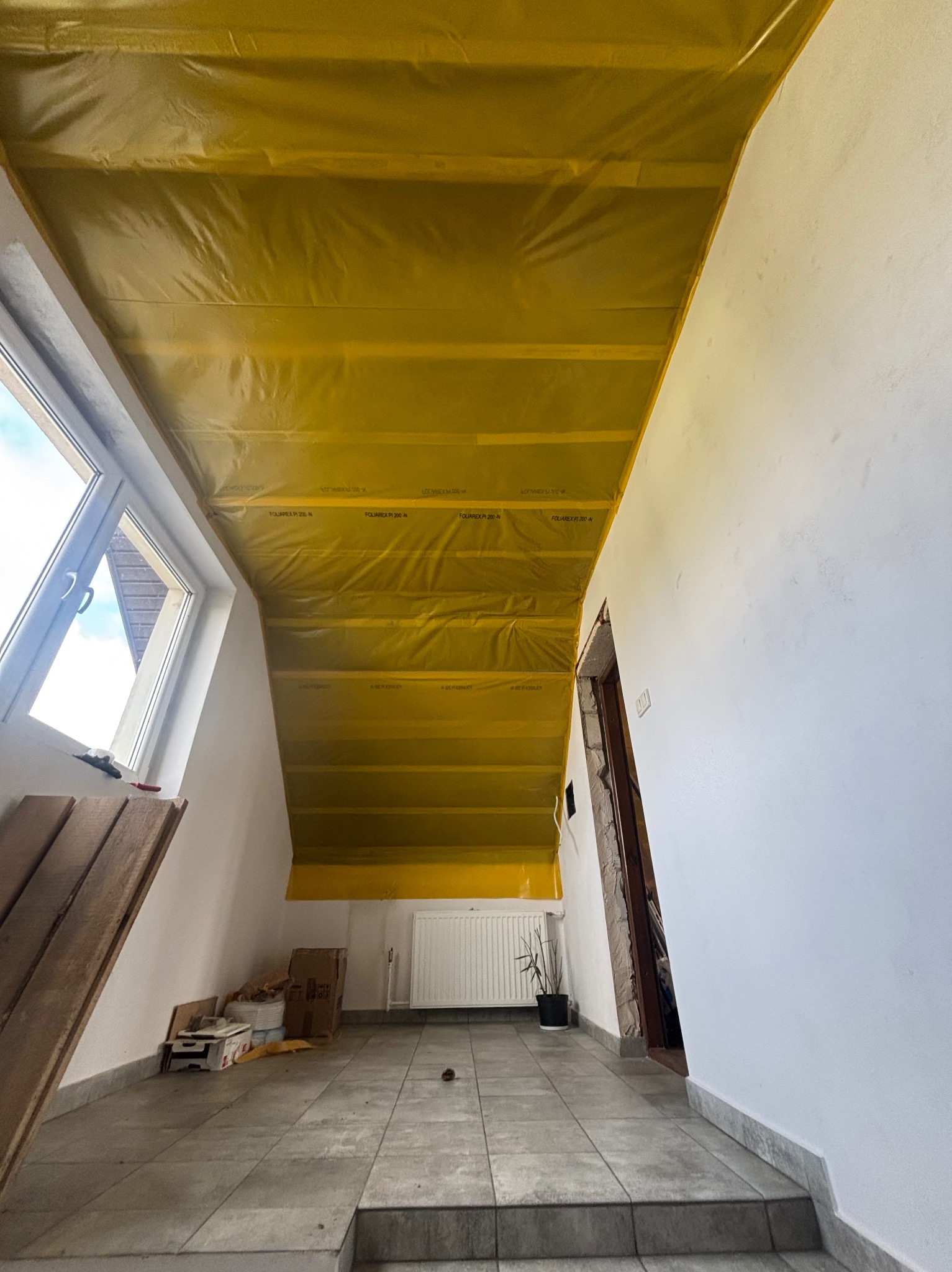 Poddasze z oknem, zabezpieczone żółtą folią paroizolacyjną na suficie. Widoczne białe ściany, grzejnik, roślina doniczkowa i płytki podłogowe w odcieniach szarości.