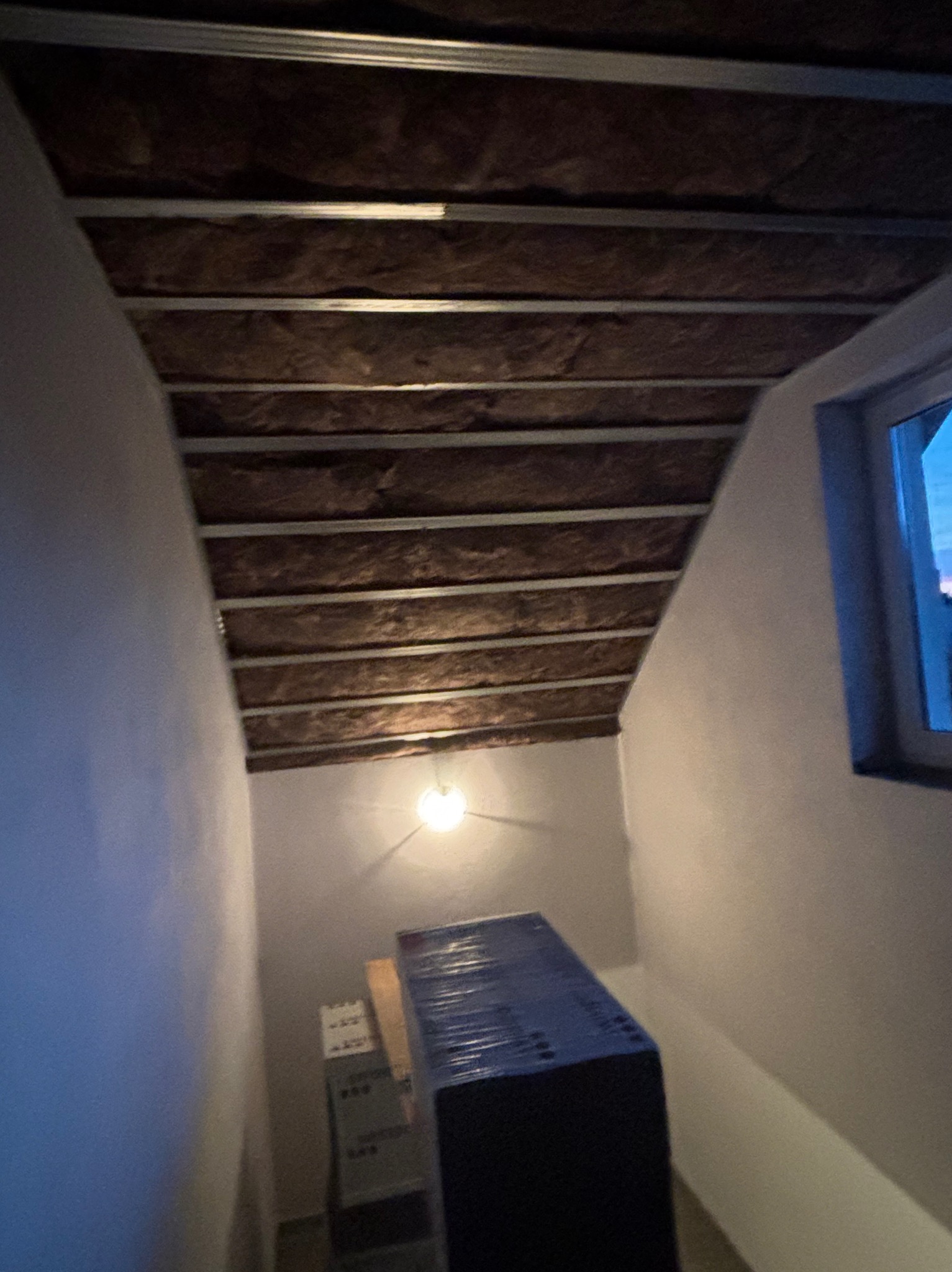 Skośny sufit poddasza z izolacją z wełny mineralnej i metalowym stelażem, widok z dołu, okno w tle. Część materiałów budowlanych na dole kadru.